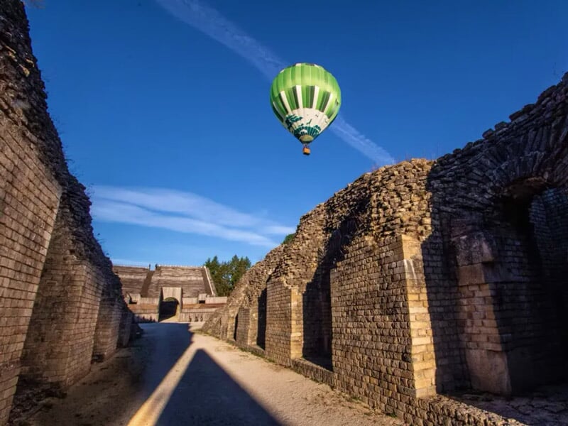 Montgolfière survolant un amphithéâtre romain avec gradins et ciel bleu.