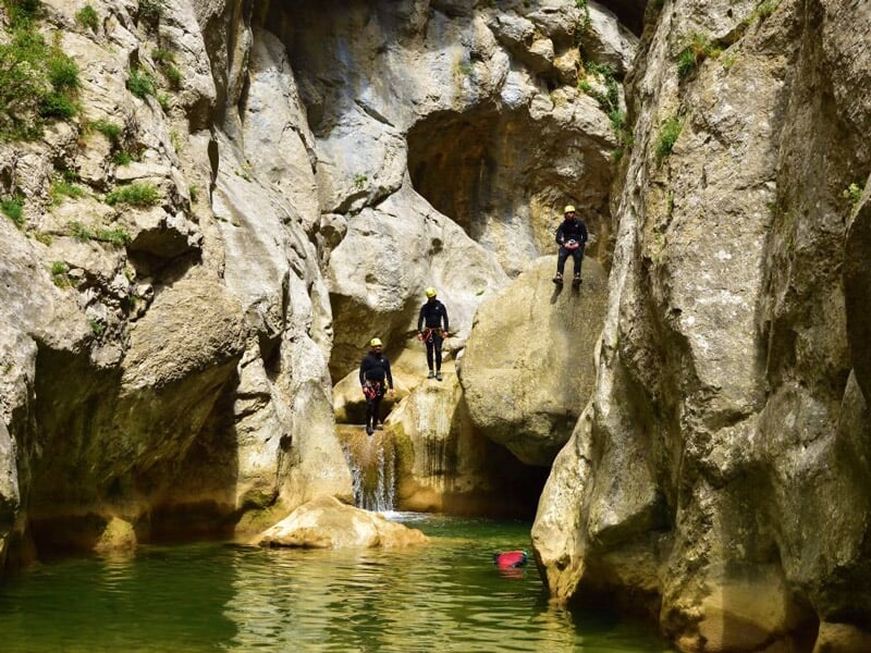 Groupe de personnes équipées de casques et combinaisons dans un canyon rocheux avec une cascade et un bassin d'eau.