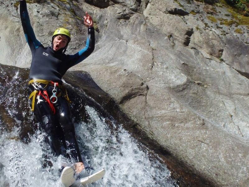 Homme en combinaison noire descend en glissade sur une cascade rocheuse avec expression joyeuse.