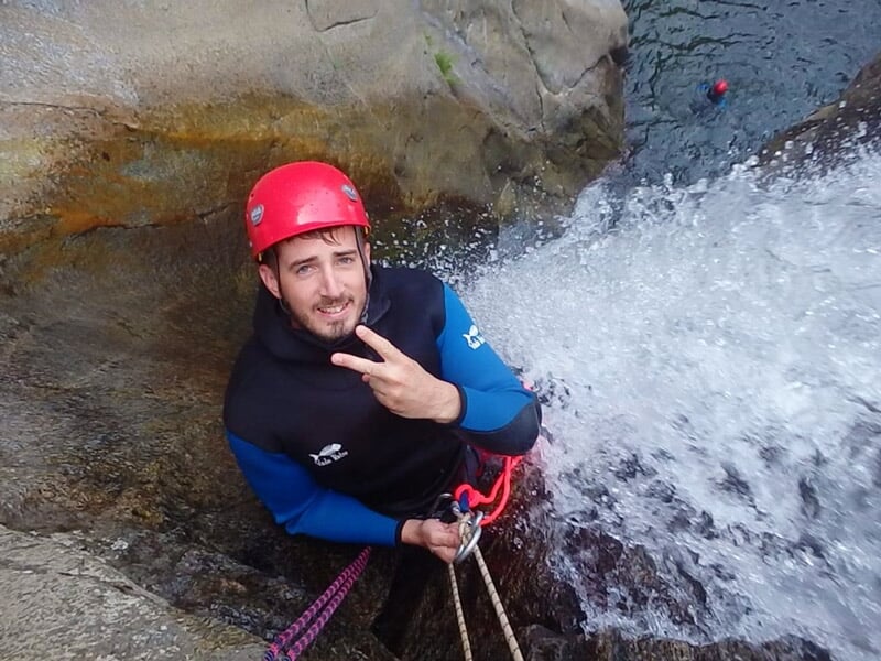 Homme en combinaison noire et casque rouge fait un signe de paix en descendant en rappel dans un canyon.