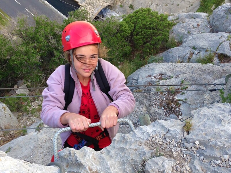 Femme en casque rouge grimpe une paroi rocheuse équipée d'une via ferrata avec un paysage en arrière-plan.