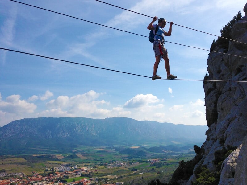 Personne en casque bleu traverse un pont de singe suspendu entre deux falaises rocheuses en via ferrata.