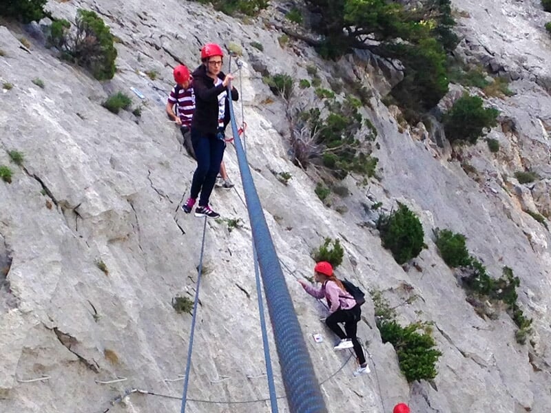Groupe de personnes en casque rouge et rose escaladent une paroi rocheuse équipée d'une via ferrata.