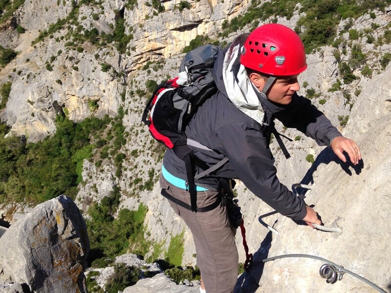 Homme en casque rouge grimpe une paroi rocheuse abrupte avec équipement de via ferrata en pleine nature.