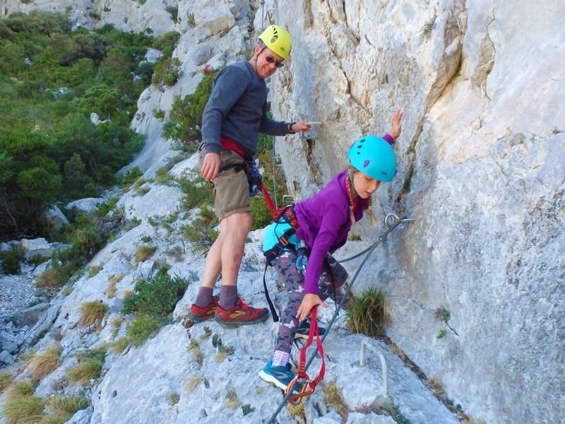 Enfant en casque bleu et combinaison violette grimpe une paroi rocheuse équipée d'une via ferrata.