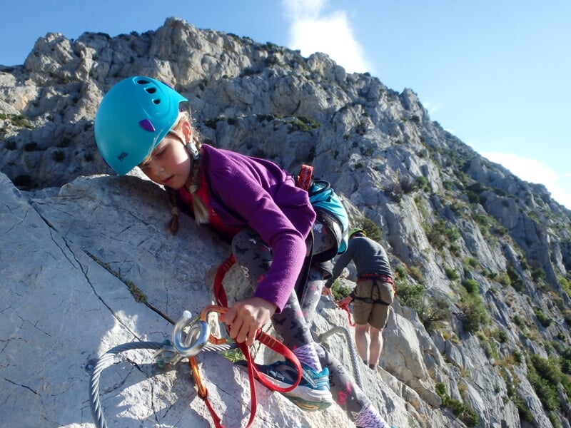 Enfant en casque bleu grimpe une paroi rocheuse avec un adulte en casque jaune en arrière-plan.