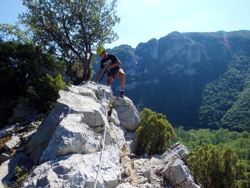 Homme en casque noir grimpe une paroi rocheuse abrupte équipée d'une via ferrata dans un paysage vert.