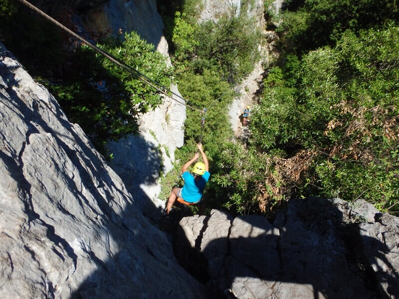 Enfant en casque bleu grimpe une paroi rocheuse équipée d'une via ferrata avec un paysage boisé.