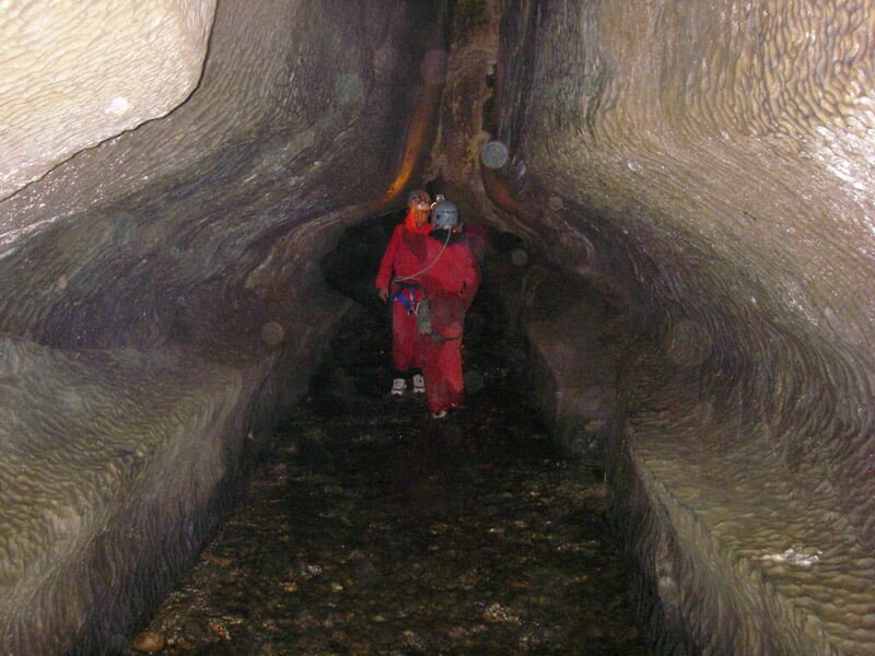 Deux spéléologues en combinaison rouge et casque avec lampe frontale explorent une galerie étroite.