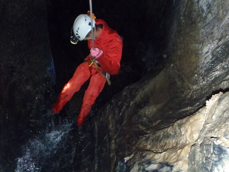 Spéléologue en combinaison rouge descend en rappel dans une cavité rocheuse humide et étroite.