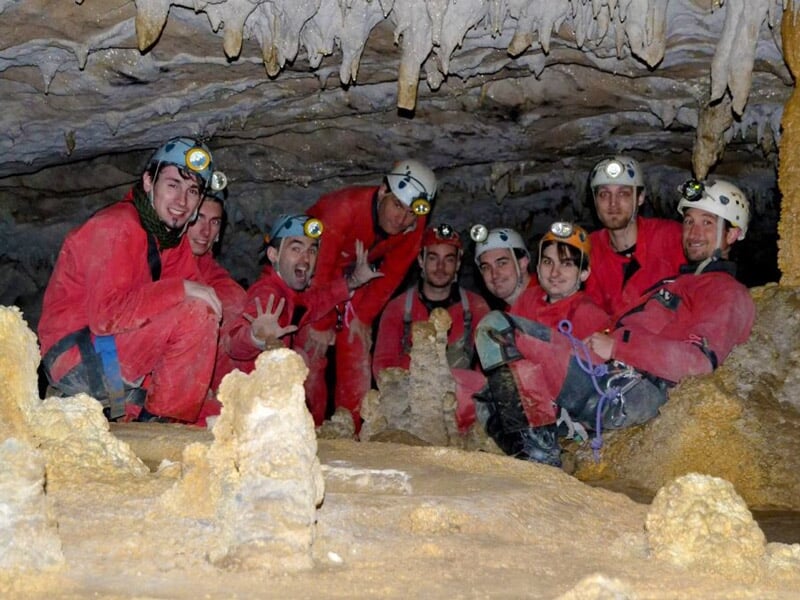 Spéléologue en combinaison rouge descend en rappel dans une cavité rocheuse sombre et humide.