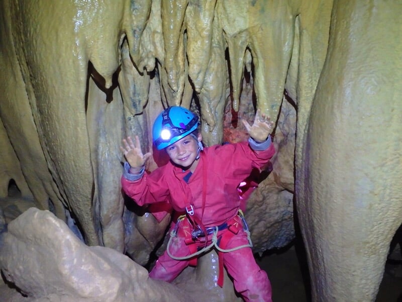 Spéléologue en combinaison rouge souriant dans une cavité rocheuse avec stalactites et stalagmites.