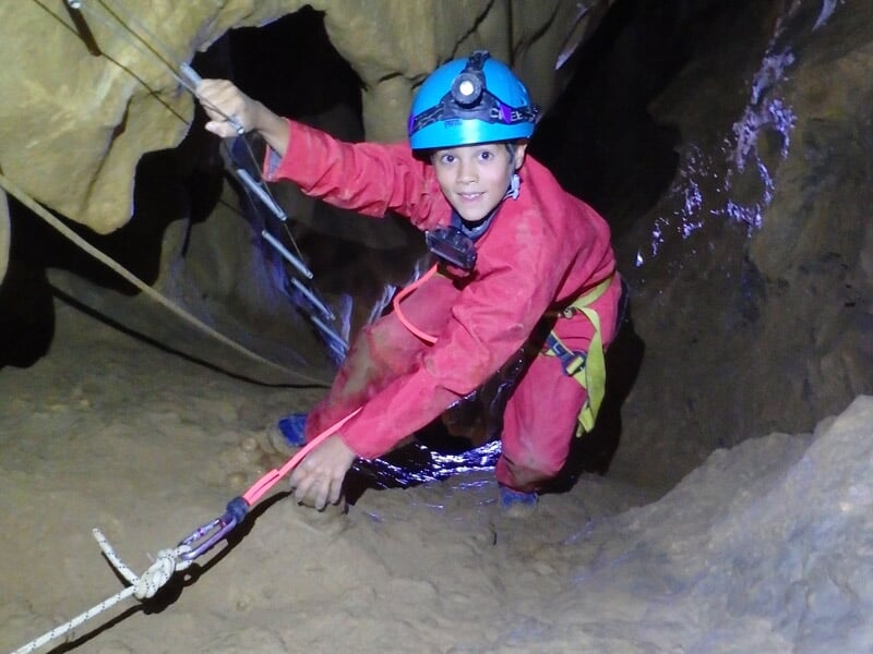 Groupe d'enfants en combinaison rouge assis dans une grotte écoutant un guide spéléologue.