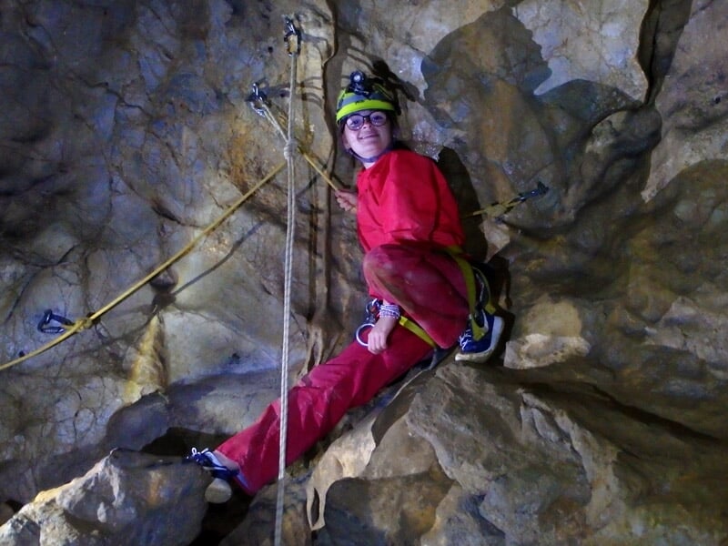 Enfant en combinaison rouge souriant dans une grotte avec stalactites et stalagmites.