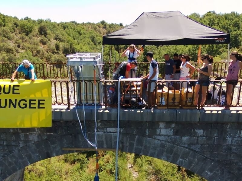 Personnes sur un pont en pierre préparant un saut à l'élastique avec une tente noire et une bannière jaune.