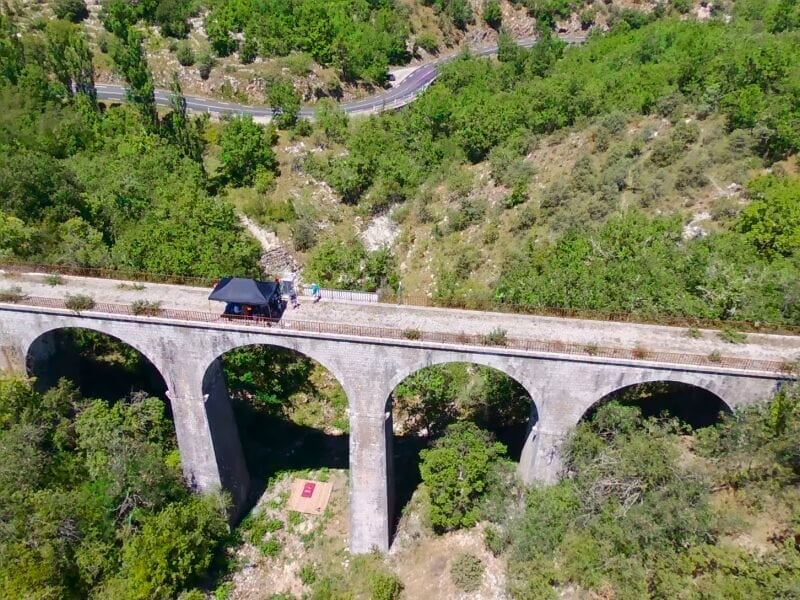 Vue aérienne d'un viaduc en pierre traversant une vallée verdoyante sous un ciel clair.