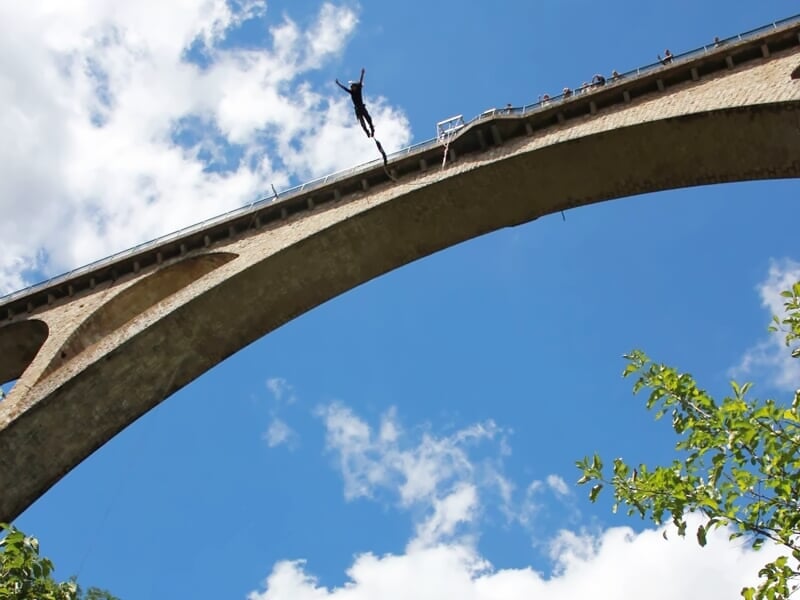 Saut à l'élastique depuis un pont en béton avec un ciel bleu et des arbres en arrière-plan.