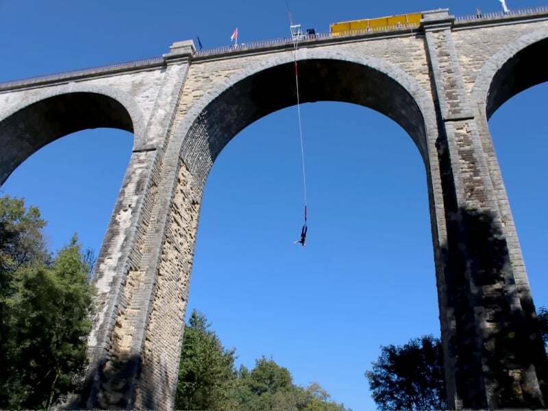 Saut à l'élastique depuis un viaduc en pierre avec un ciel bleu et une personne suspendue.