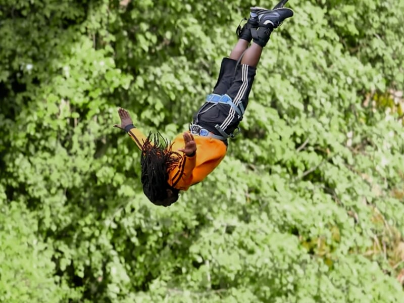 Personne en saut à l'élastique avec dreadlocks, vêtue d'un sweat orange, au-dessus des arbres.