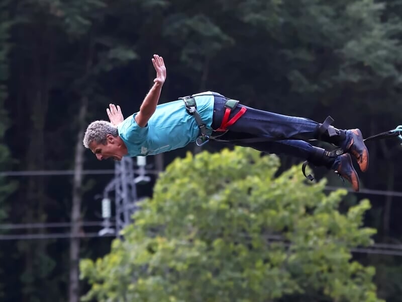 Homme en saut à l'élastique avec un t-shirt bleu clair, bras tendus, au-dessus d'une forêt.