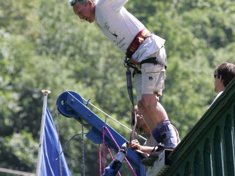 Homme en short beige sur le point de sauter à l’élastique depuis un pont, encadré par deux assistants en train de fixer les sangles.