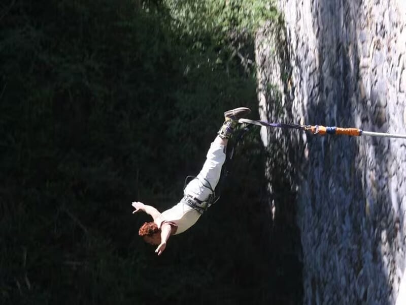 Personne en tenue blanche effectuant un saut à l’élastique, bras écartés, suspendue au-dessus d’un ravin ombragé et d’une paroi rocheuse.
