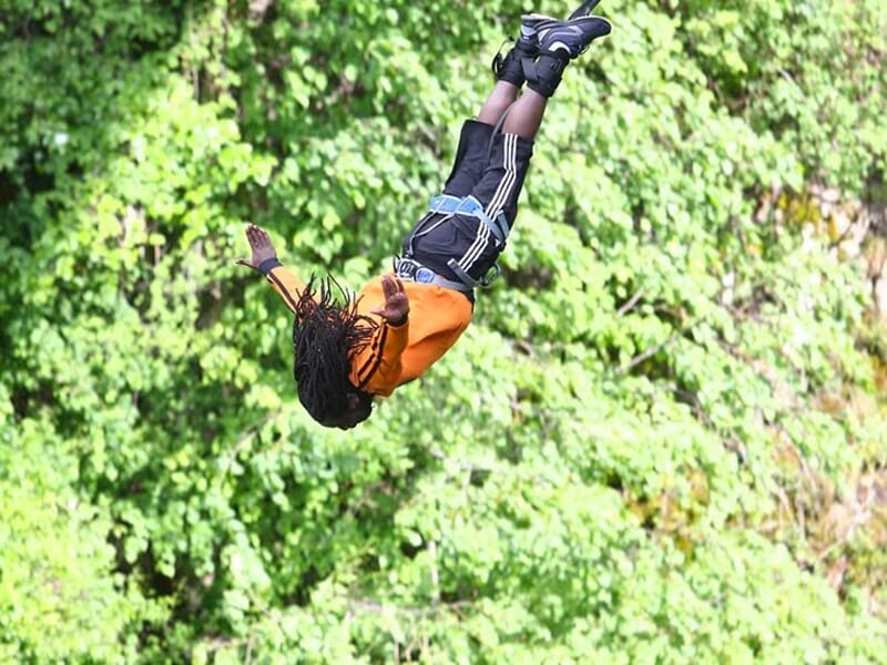 Personne en plein saut à l’élastique, torse en l’air, bras ouverts, vêtue d’un short noir et sweat orange, fond de feuillage dense.