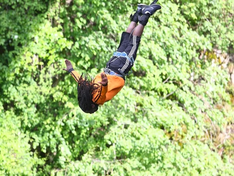 Personne en plein saut à l’élastique, torse en l’air, bras ouverts, vêtue d’un short noir et sweat orange, fond de feuillage dense.