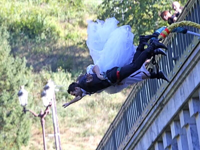 Couple en tenue de mariage effectuant un saut à l’élastique simultané depuis un pont, en position horizontale.