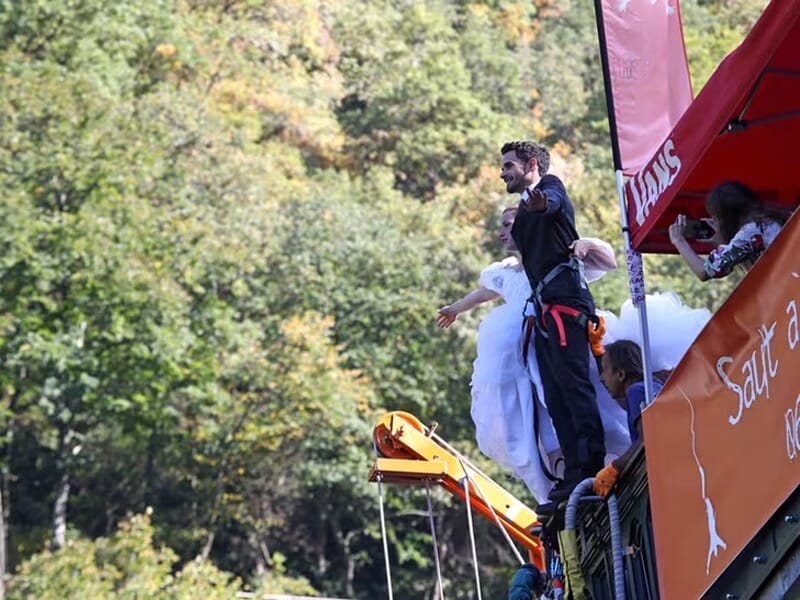 Couple en tenue de mariage debout au bord d’un pont, prêts à sauter à l’élastique, encadré par une équipe sous une tente orange.
