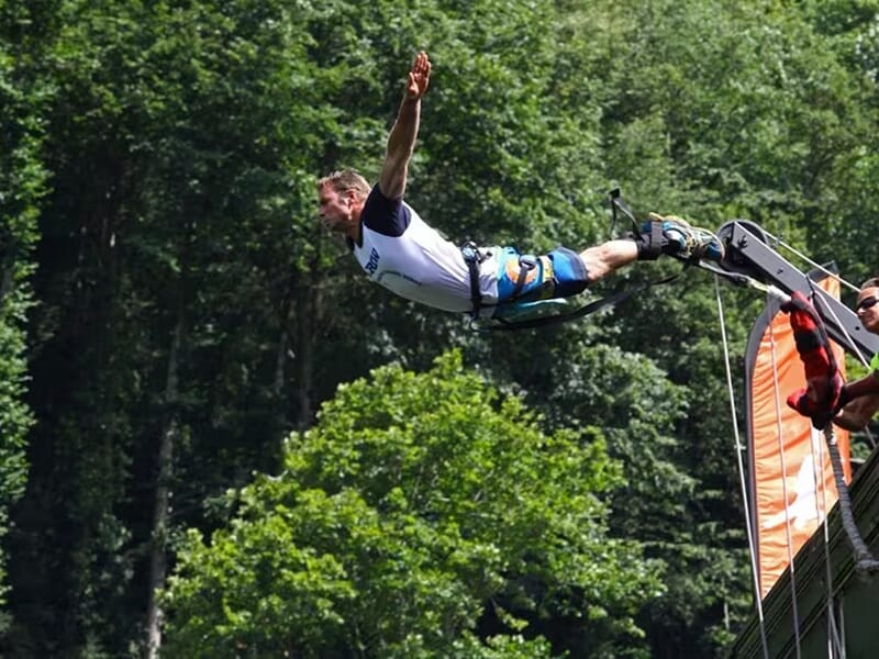 Participant en vol plané bras ouverts lors d’un saut à l’élastique, attaché par les chevilles, forêt dense en arrière-plan.