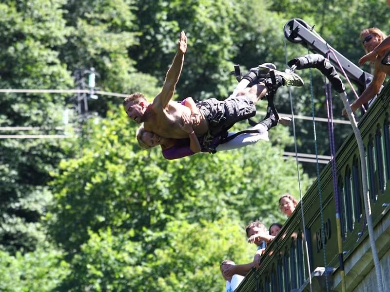 Duo en chute libre attaché ensemble pour un saut à l’élastique depuis un pont, torse nu et sourires visibles, spectateurs en arrière-plan.