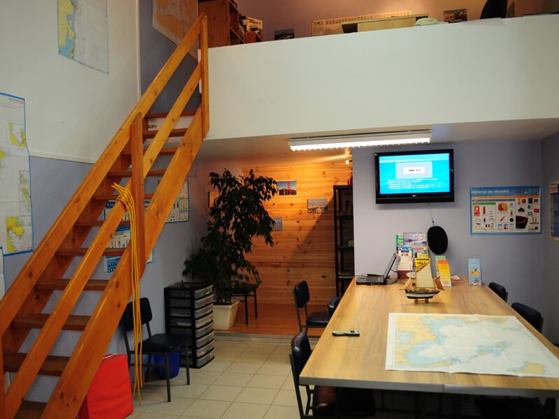 Salle de classe avec table, chaises, carte marine, écran et escalier en bois pour formation au permis bateau.