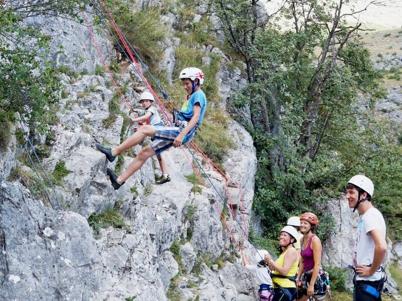 Deux enfants en casque descendent en rappel sur une paroi rocheuse avec des adultes qui les surveillent attentivement.