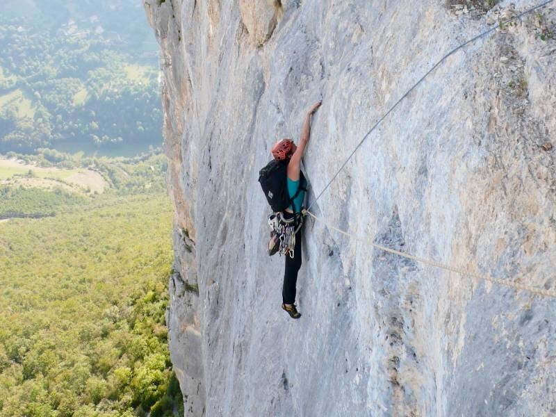 Une femme équipée d'un sac à dos grimpe une paroi rocheuse verticale avec un paysage naturel en contrebas.