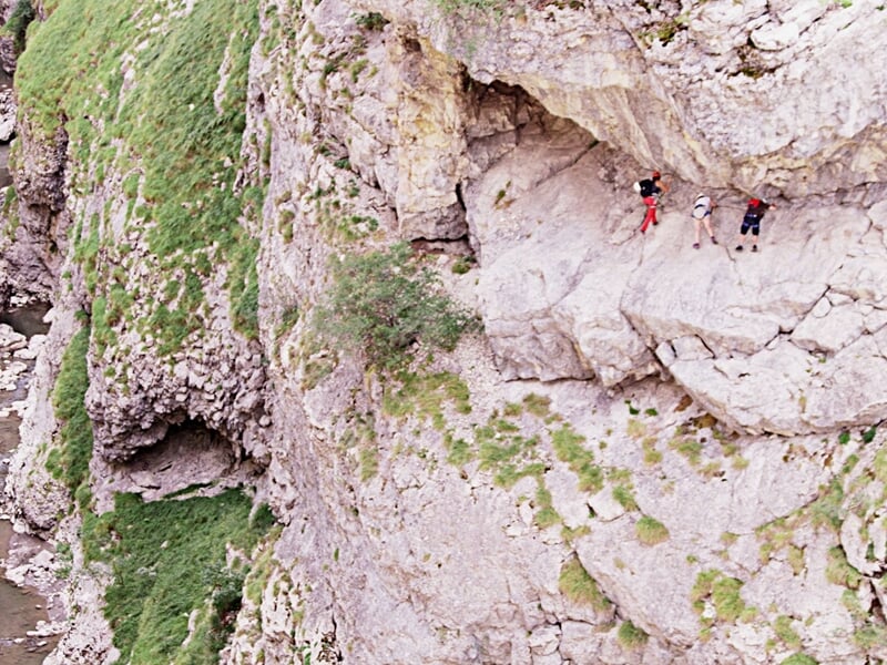 Deux personnes progressent sur un sentier étroit en falaise, entourées de rochers et de végétation.