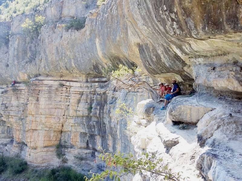 Trois personnes assises sur une corniche rocheuse surplombant une vallée, équipées pour la via ferrata.