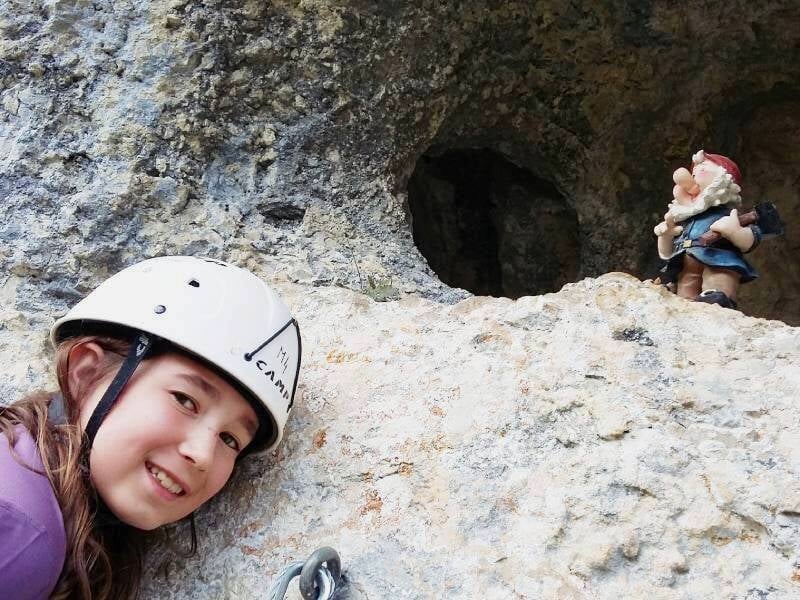 Une jeune fille en casque sourit à la caméra, posant près d'une paroi rocheuse avec une figurine de nain.