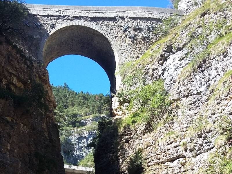 Vue d'un pont en pierre au-dessus d'une gorge rocheuse avec un ciel bleu clair en arrière-plan.