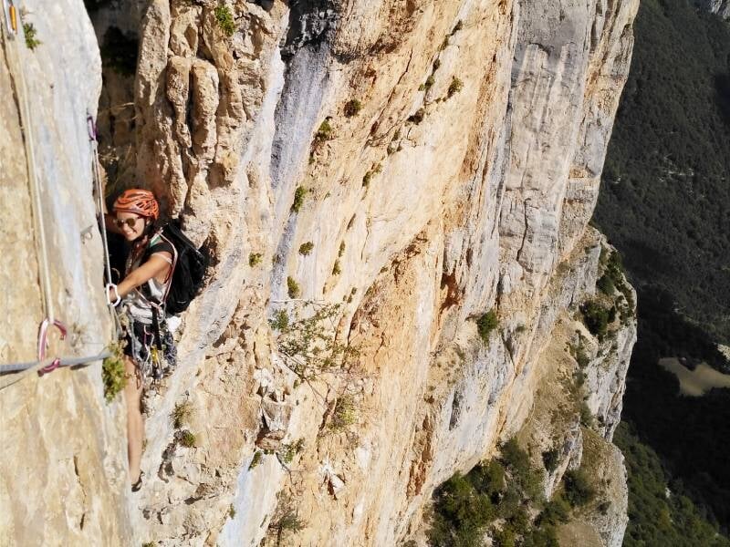 Une grimpeuse en casque escalade une paroi rocheuse abrupte avec un paysage montagneux en arrière-plan.