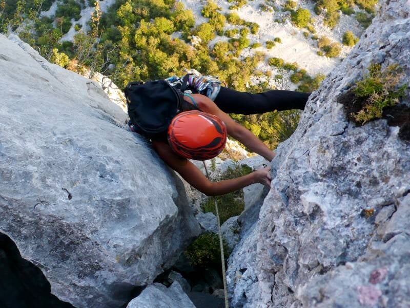 Une personne équipée d'un casque et d'un sac à dos grimpe entre deux rochers sur une paroi escarpée.