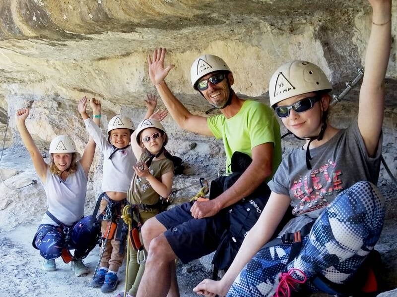 Un groupe de cinq personnes en casque et équipement de via ferrata pose joyeusement sous une corniche rocheuse.