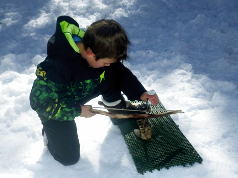 Enfant en tenue d'hiver préparant des raquettes à neige sur un tapis vert dans la neige blanche éclatante.