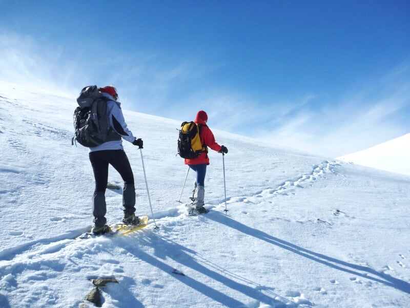 Deux randonneurs en raquettes à neige marchant sur un sentier enneigé sous un ciel bleu clair lumineux.