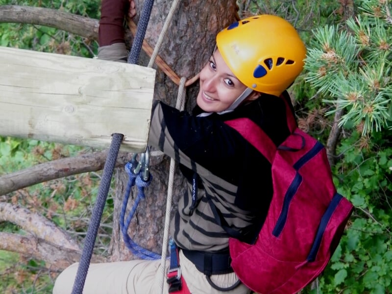 Jeune femme souriante équipée d'un casque jaune et harnais, escaladant une via ferrata en forêt verdoyante.