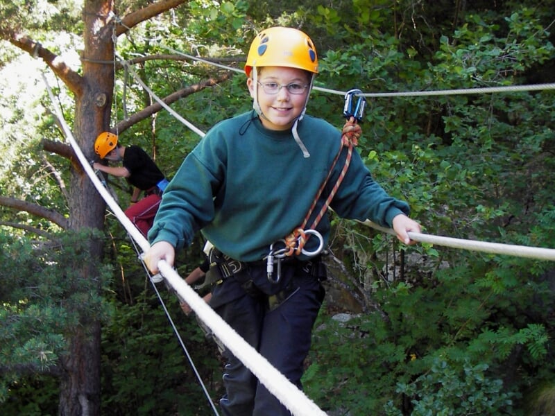 Enfant en casque jaune franchissant un pont de corde suspendu dans un parcours d'aventure en forêt verdoyante.