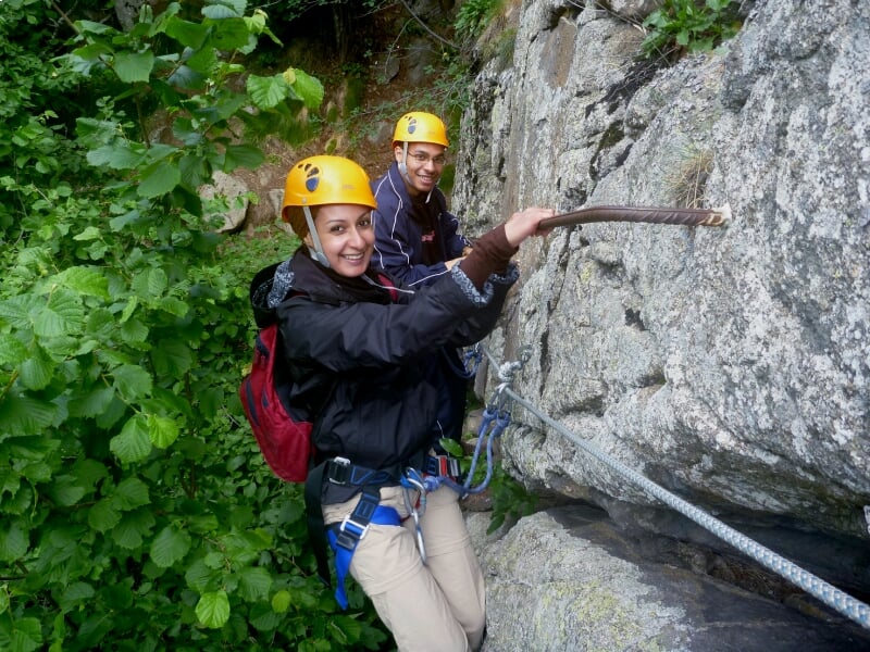 Couple équipé de casque et harnais progressant sur une via ferrata rocheuse entourée de végétation dense.