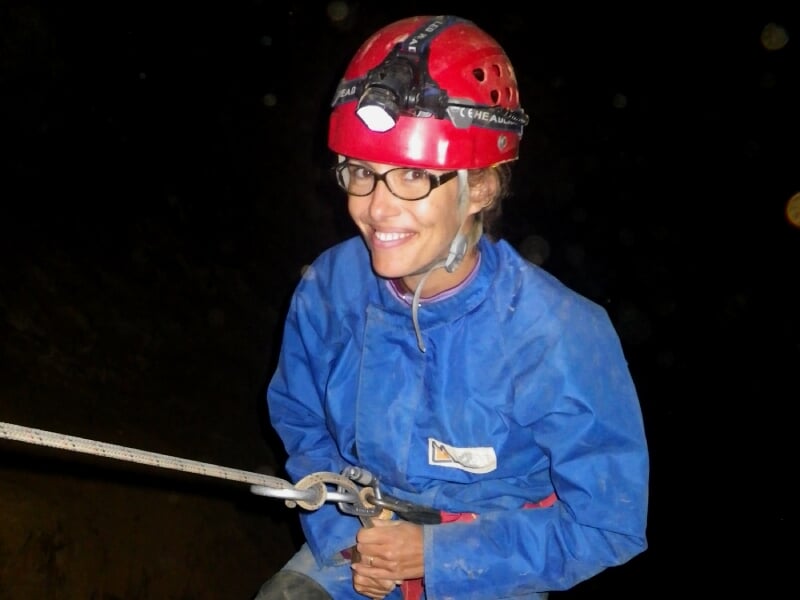 Femme souriante en combinaison bleue et casque rouge avec lampe frontale, explorant une grotte sombre en spéléologie.