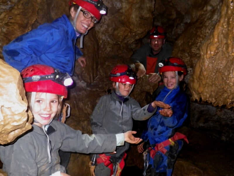 Groupe d'enfants en combinaison et casque rouge avec lampe frontale, explorant une cavité rocheuse en spéléologie.