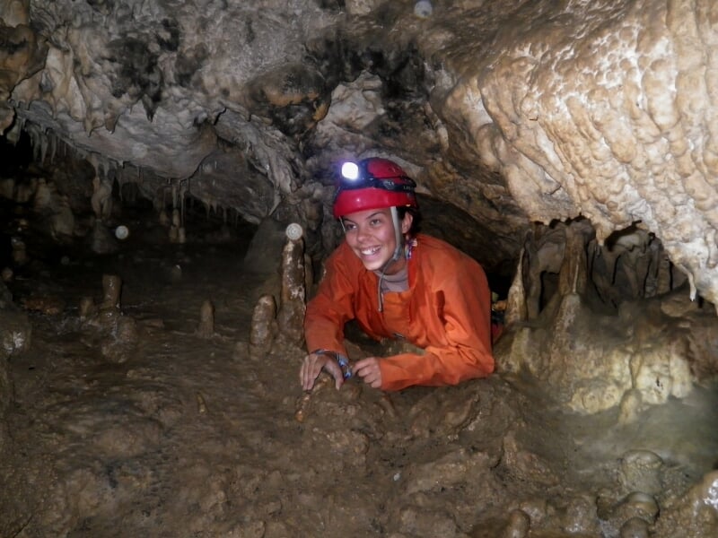 Personne en combinaison orange et casque rouge avec lampe frontale, souriant dans une grotte aux formations rocheuses.